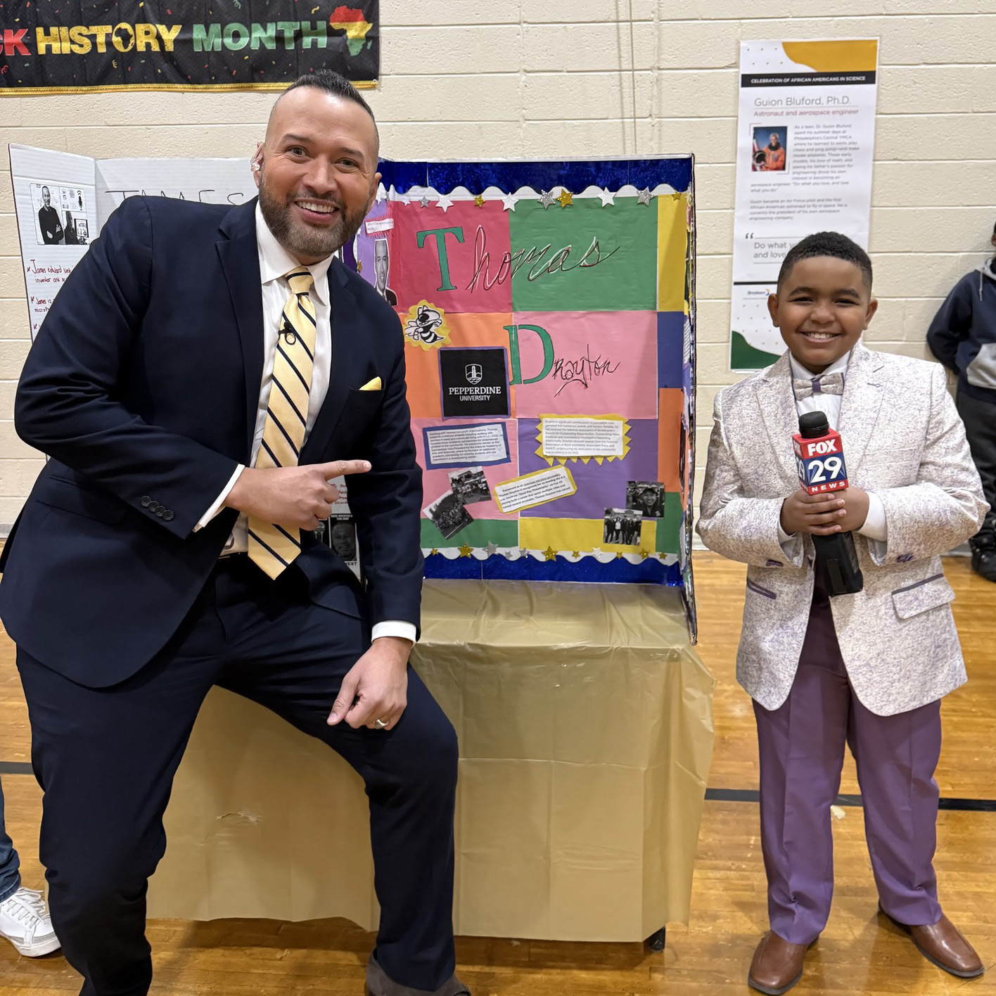 Student dressed as a news reporter holding a FOX 29 microphone stands beside a presenter during a Black History Month project showcase at Edward Heston School in Philadelphia.